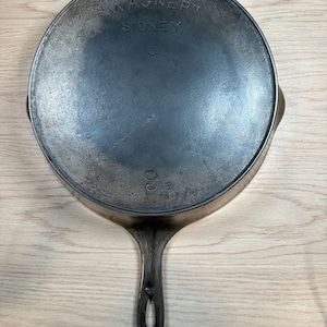 May include: A round cast iron skillet with a long handle featuring a central hole. The underside is embossed with the text WAGNER SIDNEY O. and the number 9 B. The metal surface shows a dark, seasoned patina with visible texture and wear.