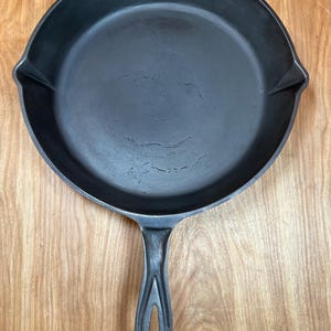 May include: A black cast iron skillet with a long handle and a rounded cooking surface. The skillet has a small spout on either side of the rim. The handle has a hole at the end for hanging.