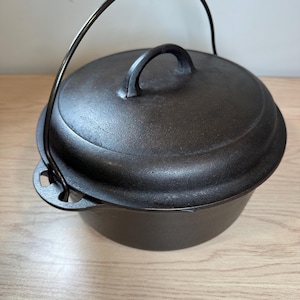 May include: A round, black cast iron Dutch oven with a matching lid and a curved metal bail handle. The pot features two small side handles integrated into the main body, and the lid has a central handle for lifting. The surface is dark.