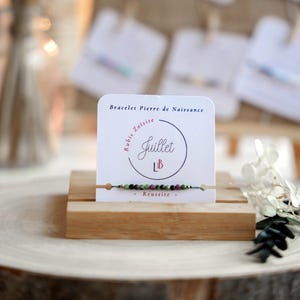 Peut inclure: Un bracelet avec des perles vertes et roses sur un support en bois. La carte indique "Bracelet Pierre de Naissance", "Rubis Zoisite", "Juillet", "Lib" et "Reussite".