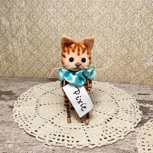 Może przedstawiać: Ręcznie wykonana filcowa figurka kota z pomarańczowymi i brązowymi paskami, nosząca jasnoniebieską wstążkę. Dołączona jest etykieta z napisem "Pixie". Kot stoi na białej koronkowej serwetce.
