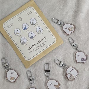 Peut inclure: Plusieurs porte-clés mystères Little Escapes en acrylique, représentant un personnage fantomatique avec divers accessoires. Les porte-clés mesurent 3,8 cm et sont dotés de fermoirs argentés. Une carte avec le design est également présente.