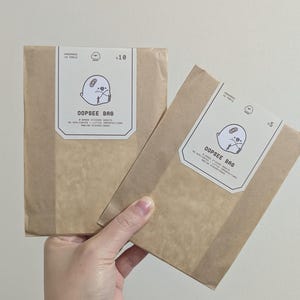 Peut inclure: Deux paquets en papier brun sont tenus par une main. Ils portent des étiquettes blanches avec le texte "OOPSSEE BAG" et un dessin de phoque. L'un indique "x10", l'autre "x5". Les paquets semblent contenir des autocollants.