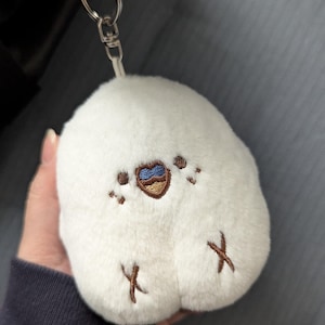 Peut inclure: Un porte-clés en peluche blanc en forme d'oiseau. L'oiseau a un motif en forme de cœur marron et bleu sur sa poitrine, avec des détails brodés. Il est attaché à un anneau en métal argenté.