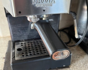 May include: A black espresso machine with a silver top. The machine has a silver handle with the word "Gaggia" and "Classic" printed on it. The machine is on a black surface.