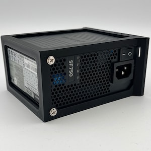 May include: A black SF750 power supply unit with a honeycomb grill and an on/off switch. The unit has a power input port and is secured with silver screws. The SF750 model name is visible on the side.