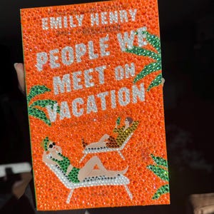 May include: An orange book cover with the title "People We Meet on Vacation" by Emily Henry. The cover is decorated with small, sparkling orange and silver accents. Illustrations of people relaxing on sun loungers are featured.