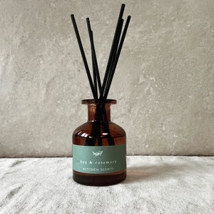May include: A brown glass diffuser bottle with black reeds. The bottle has a green label with the text "bay & rosemary KITCHEN SCENTS". The diffuser is designed to add fragrance to a kitchen or living space.