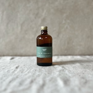 Könnte beinhalten: Eine braune Glasflasche mit goldenem Deckel und einem grünen Etikett mit der Aufschrift "bay & rosemary" und "KITCHEN SCENTS". Die Flasche ist mit einer klaren Flüssigkeit gefüllt. Der Hintergrund ist eine neutrale, strukturierte Oberfläche.
