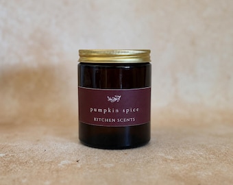 May include: A dark amber glass candle jar with a gold-colored metal lid. The jar features a maroon label with a small white leaf icon, the text "pumpkin spice" in lowercase, and the text "KITCHEN SCENTS" in uppercase letters centered on the front.