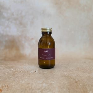 Könnte beinhalten: Eine kleine bernsteinfarbene Glasflasche mit einem goldfarbenen Metallschraubverschluss. Die Flasche hat ein dunkelrotes, rechteckiges Etikett mit einer weißen botanischen Illustration und dem Text "fireside KITCHEN SCENTS" in weißer Schrift.