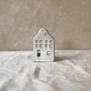Puede incluir: Una pequeña figura de casa de cerámica blanca con un detalle en forma de corazón en la parte superior. La casa presenta múltiples ventanas cuadradas y una puerta. La casa en miniatura es un objeto decorativo.