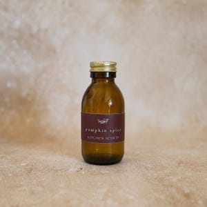 May include: A small amber glass bottle with a gold screw-top lid. A dark maroon label is wrapped around the center, featuring a white line drawing of a sprig and the text "pumpkin spice" and "KITCHEN SCENTS" printed in white lettering.