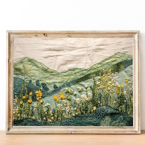 May include: Embroidered landscape artwork in a wooden frame. The textile art features green hills and a meadow of yellow and white flowers. The frame is light-coloured, adding to the rustic charm. The piece is likely handmade, with detailed embroidery.