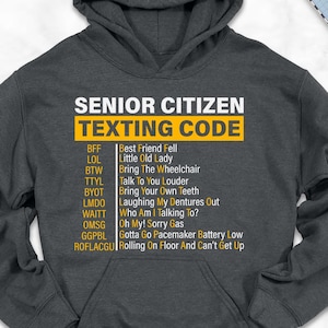 May include: A dark gray hoodie with the text "SENIOR CITIZEN TEXTING CODE" in yellow and white. The hoodie is surrounded by a scarf, shoes, jeans, and a backpack.