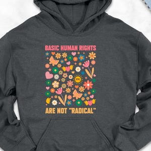 May include: A dark gray hoodie with the text "BASIC HUMAN RIGHTS ARE NOT 'RADICAL'" in pink and yellow. The design includes colorful flowers, butterflies, and hearts. The hoodie is surrounded by accessories like shoes, jeans, and a backpack.
