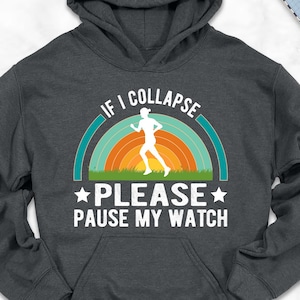 May include: Dark gray hoodie with the text "IF I COLLAPSE PLEASE PAUSE MY WATCH" above a running figure inside a rainbow. The hoodie is surrounded by casual items like shoes, jeans, and a backpack.