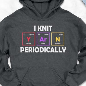 May include: Dark gray hoodie with the text "I KNIT YARN PERIODICALLY" in white. The design incorporates the periodic table elements for Yarn. The hoodie has a front pocket and a hood. The image also shows jeans, shoes, and a scarf.