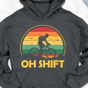 May include: Dark gray hoodie with a cycling graphic inside a sunset design. The hoodie features the text "OH SHIFT" in yellow. The image includes a scarf, shoes, jeans, and other accessories.