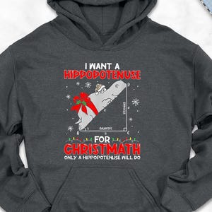 May include: Dark gray hoodie with the text "I WANT A HIPPOPOTENUSE FOR CHRISTMATH ONLY A HIPPOPOTENUSE WILL DO." The graphic includes a Christmas bow and a right triangle. The hoodie is surrounded by accessories.