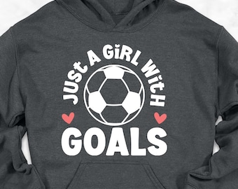 Könnte beinhalten: Ein anthrazitfarbener Kapuzenpullover mit dem Text "JUST A GIRL WITH GOALS" in Weiß und einer Fußballgrafik. Der Kapuzenpullover hat eine Kapuze mit Kordelzug und eine Vordertasche. Das Bild enthält Jeans, Schuhe und einen Rucksack.