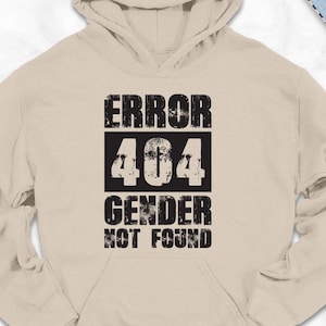 May include: A beige hoodie with the text "ERROR 404 GENDER NOT FOUND" in a black, distressed font. The hoodie is laid flat, surrounded by accessories like shoes, a scarf, and a backpack. The hoodie is a casual garment.