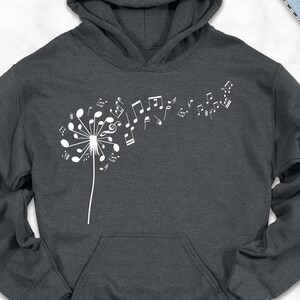 May include: A dark gray hoodie with a white dandelion design composed of musical notes. The hoodie has a front pocket and a hood. The image also includes a pair of brown shoes, blue jeans, and a scarf.