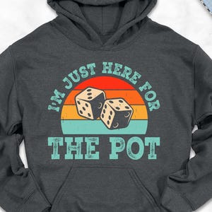 May include: Dark gray hoodie with the text "I'M JUST HERE FOR THE POT" and dice graphic. The hoodie has a drawstring hood and a front pocket. The image also includes a pair of brown shoes, jeans, and a scarf.