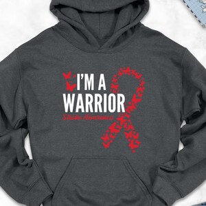May include: A dark gray hoodie with the text "I'M A WARRIOR" in white and red. The design includes a red ribbon with butterfly accents. The hoodie is surrounded by accessories like shoes, jeans, and a scarf.