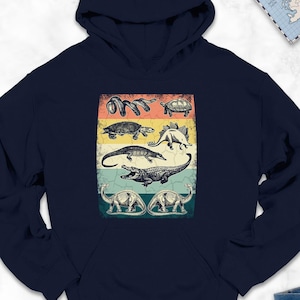 May include: Navy blue hoodie featuring a vintage-style graphic of various prehistoric animals, including dinosaurs and other reptiles, arranged in a vertical rectangular design. The graphic has a retro color scheme with a distressed look. The hoodie has a hood and a kangaroo pocket.