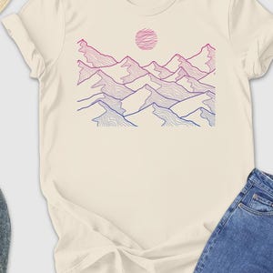 May include: Cream-colored t-shirt featuring a mountain range design in shades of pink, purple, and blue, with a matching pink sun. The shirt is paired with blue jeans, gray shoes, a straw hat, sunglasses, and a yellow wallet.