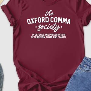 May include: A maroon t-shirt with the text "the OXFORD COMMA society" in white, along with the phrase "IN DEFENSE AND PRESERVATION OF TRADITION, FORM, AND CLARITY." The shirt is paired with blue jeans, gray shoes, and a straw hat.