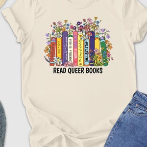 May include: Cream-colored t-shirt featuring a colorful graphic of books with floral accents and the text "READ QUEER BOOKS." The shirt is paired with blue jeans, gray sneakers, and a straw hat with sunglasses.
