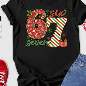 May include: Black t-shirt with a festive "67 seven" design. The "6" is red with Christmas ornaments, and the "7" is red and green striped. The shirt is surrounded by Christmas-themed items, including a Santa hat, red shoes, and gloves.