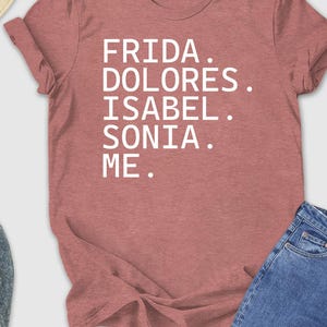 May include: A dusty rose t-shirt with the words "FRIDA. DOLORES. ISABEL. SONIA. ME." in white. The shirt is laid out with blue jeans, a straw hat, sunglasses, and sneakers.