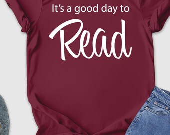 May include: A maroon t-shirt with the text "It's a good day to Read" in white script. The image also includes blue jeans, gray shoes, a straw hat, sunglasses, and a small camera.
