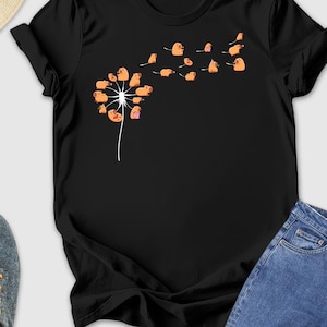 May include: Black t-shirt featuring a dandelion design with orange and pink seed heads blowing in the wind. The shirt is styled with blue jeans, gray sneakers, a straw hat, sunglasses, and a yellow wallet.