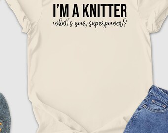 May include: A cream-colored t-shirt with the text "I'M A KNITTER what's your superpower?" in black. The image includes blue jeans, gray shoes with yellow laces, a straw hat, sunglasses, a yellow wallet, a camera, and a small potted plant.