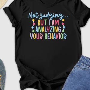 May include: Black t-shirt with the text "Not judging... BUT I AM ANALYZING YOUR BEHAVIOR" in colorful letters. The shirt is paired with blue jeans, gray sneakers, and a straw hat with sunglasses.