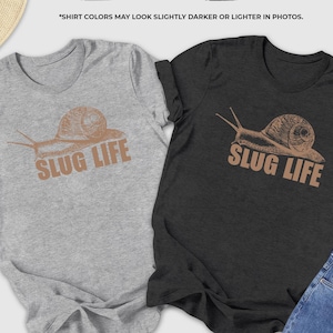 May include: Two t-shirts, one athletic heather and one dark grey heather, each with a snail graphic and the words "SLUG LIFE" in a brown font. The shirts are displayed with jeans, shoes, and accessories.