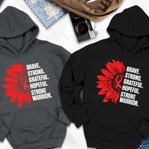 May include: Two hooded sweatshirts, one charcoal gray and one black, are displayed. Each features a red sunflower design with a stroke awareness ribbon and the words "BRAVE. STRONG. GRATEFUL. HOPEFUL. STROKE WARRIOR." The sweatshirts are on a white surface.
