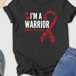 May include: Dark gray t-shirt with the text "I'M A WARRIOR" and a red ribbon made of butterflies, promoting stroke awareness. The shirt is paired with blue jeans, gray sneakers, and a straw hat with sunglasses.