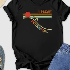 May include: Black t-shirt with the text "I HAVE POTENTIAL" in a retro design. The shirt features a red circle, horizontal stripes, and an arrow pointing down. The shirt is paired with blue jeans, sneakers, and a straw hat.