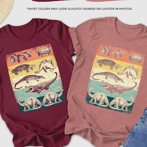 May include: Two t-shirts, one maroon and one heather mauve, each with a vintage-style dinosaur graphic. The graphic features various dinosaurs and reptiles in a colorful, retro design. The text "SHIRT COLORS MAY LOOK SLIGHTLY DARKER OR LIGHTER IN PHOTOS" is also visible.