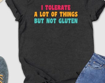 May include: Dark gray t-shirt with the text "I TOLERATE A LOT OF THINGS BUT NOT GLUTEN" in pink, yellow, and blue. The shirt is paired with blue jeans, gray sneakers, and a straw hat with sunglasses.