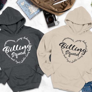 May include: Two hooded sweatshirts, one charcoal gray and one beige, with the words "Billing Squad" in a heart-shaped design. The design includes words like "compassionate" and "caring". The sweatshirts have a front pocket and a hood.