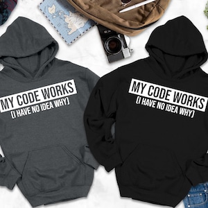 May include: Two hooded sweatshirts, one charcoal gray and one black, are displayed. Each features the text "MY CODE WORKS (I HAVE NO IDEA WHY)" in a white rectangular box. The sweatshirts have a front pocket and a hood. The bottom of the image has the text "HOODIE COLORS MAY LOOK SLIGHTLY DARKER OR LIGHTER IN PHOTOS."