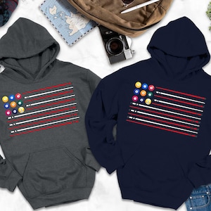 May include: Two hooded sweatshirts, one charcoal gray and one navy blue, each featuring a graphic of a billiards-themed flag. The design includes a cluster of billiard balls in the upper left corner and red and white stripes. The sweatshirts are displayed with accessories.
