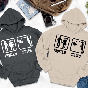 May include: Two hooded sweatshirts, one charcoal gray and one beige, with the text "PROBLEM SOLVED". Each hoodie features a graphic of a stick figure couple and a person throwing a laptop. The text "HOODIE COLORS MAY LOOK SLIGHTLY DARKER OR LIGHTER IN PHOTOS." is at the bottom.