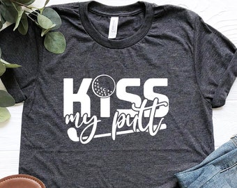May include: Dark gray t-shirt with the text "KISS my putt" in white, featuring a golf ball graphic. The shirt is paired with blue jeans and black sandals with brown soles. A sprig of greenery is also present.